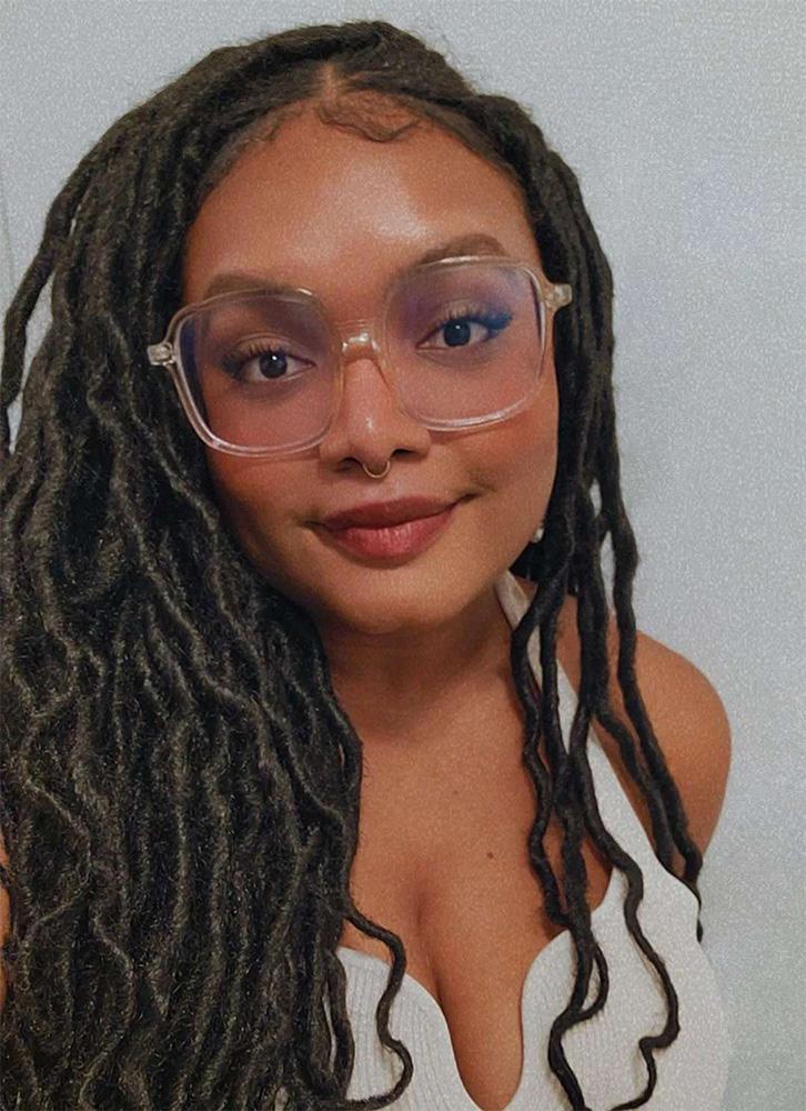 Headshot of woman with long black twists, big rimmed clear glasses, brown skin, a white tank top.