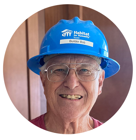 Bob is wearing a blue Habitat hard hat and glasses and he smiles for the camera. Behind him are brown kitchen cabinets. 