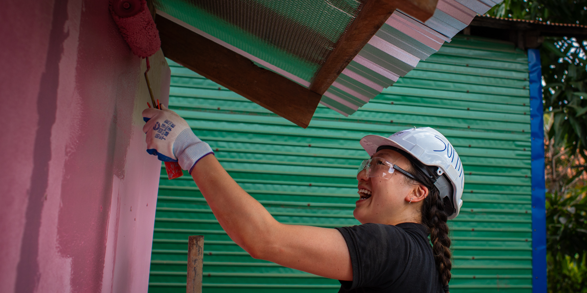 A volunteer in a white hard hat and black shirt uses a roller to paint the side of a building pink. The tin roof of the building is visible above her, and the background is filled by a vibrant green wall.  