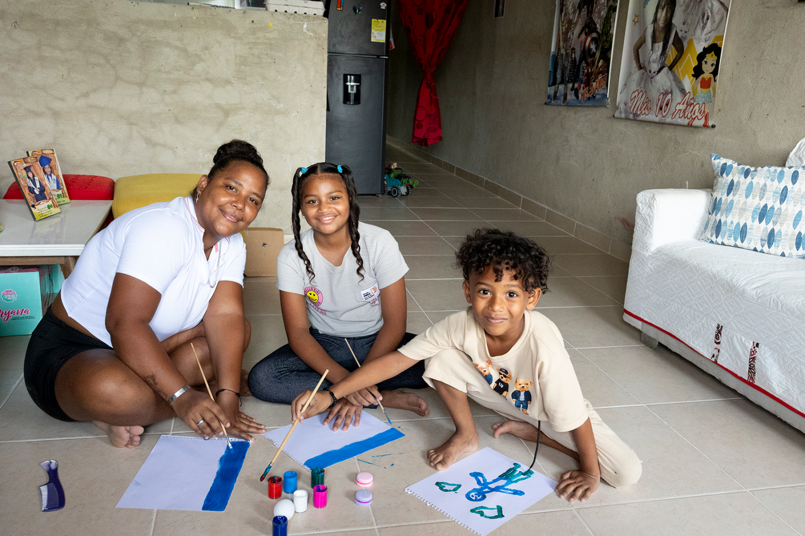 A woman sits on a tile floor with her two kids painting on white sheets of paper with blue paint.