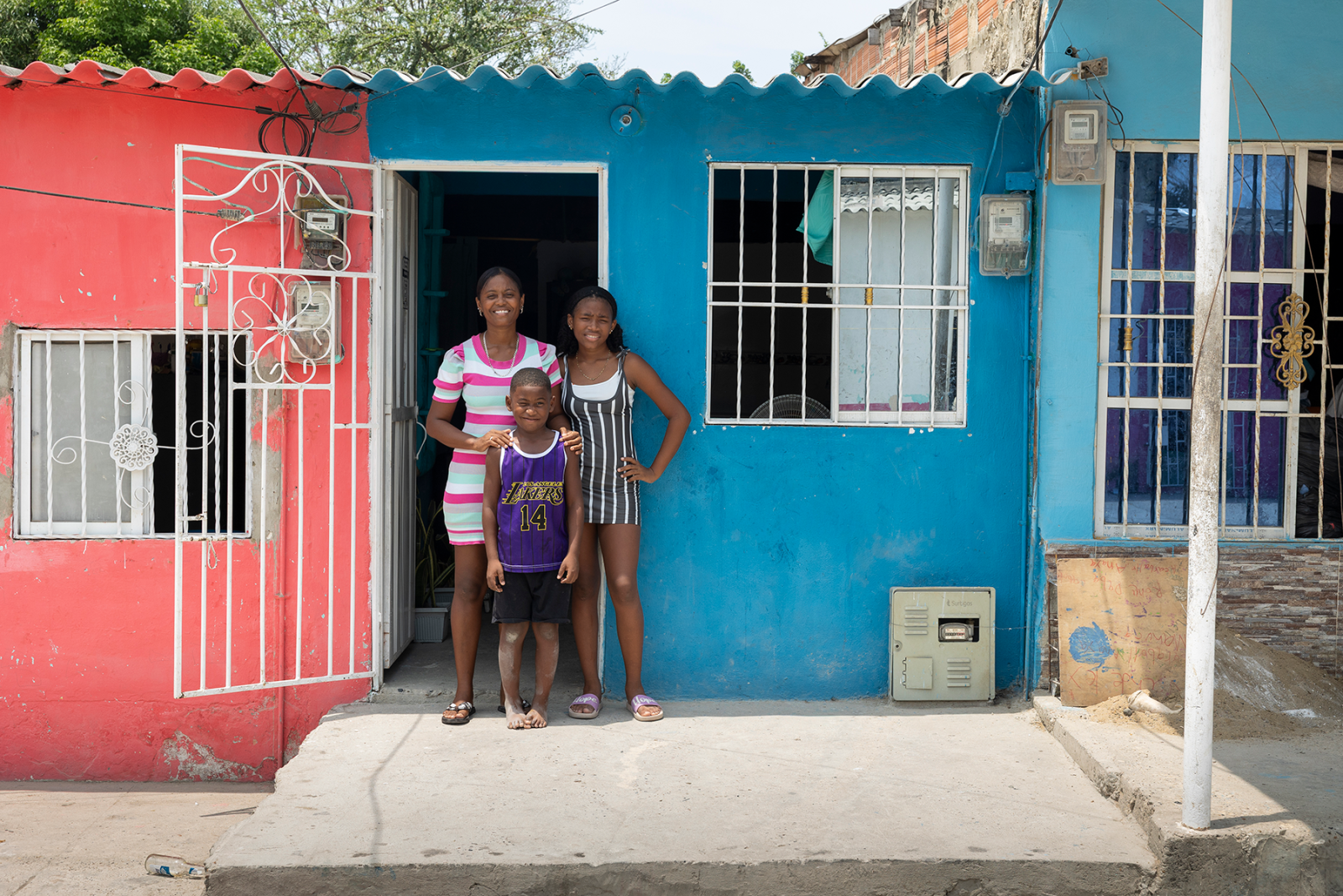 Una mujer está de pie con sus dos hijos frente a su casa, que está pintada de color melocotón y azul. Hay un porche de cemento a sus pies.