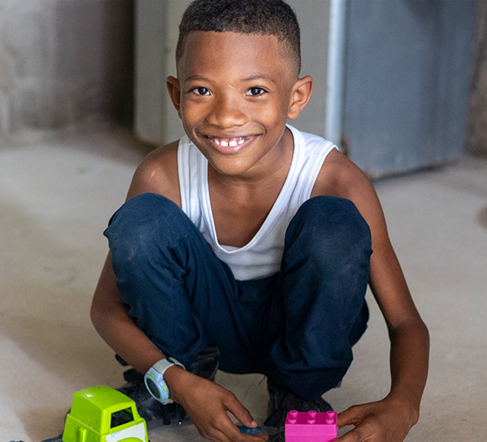A child wearing jeans and a tank top squats on the clean floor to play with a green toy car and pink lego.