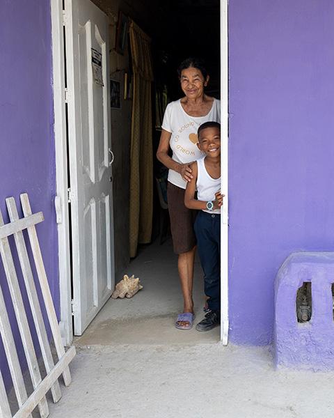 An older woman and a young boy peek out from the doorway of a bright lilac house with clean-swept concrete flooring.