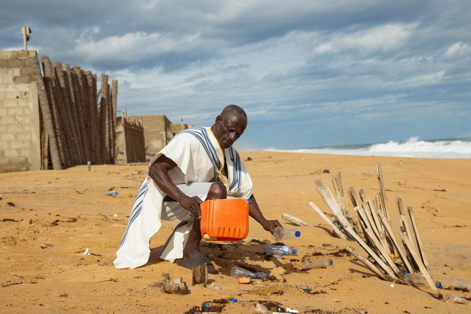 A man named Alex dressed in white crouches down and holds an orange bucket and collects plastic bottles off the beach near his home.