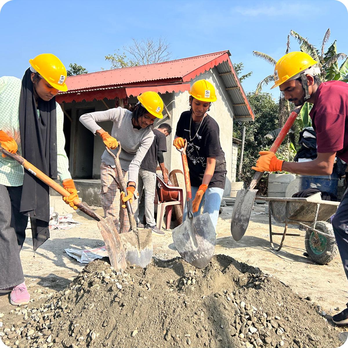 Four young people in yellow hardhats shoveling a pile of dirt.