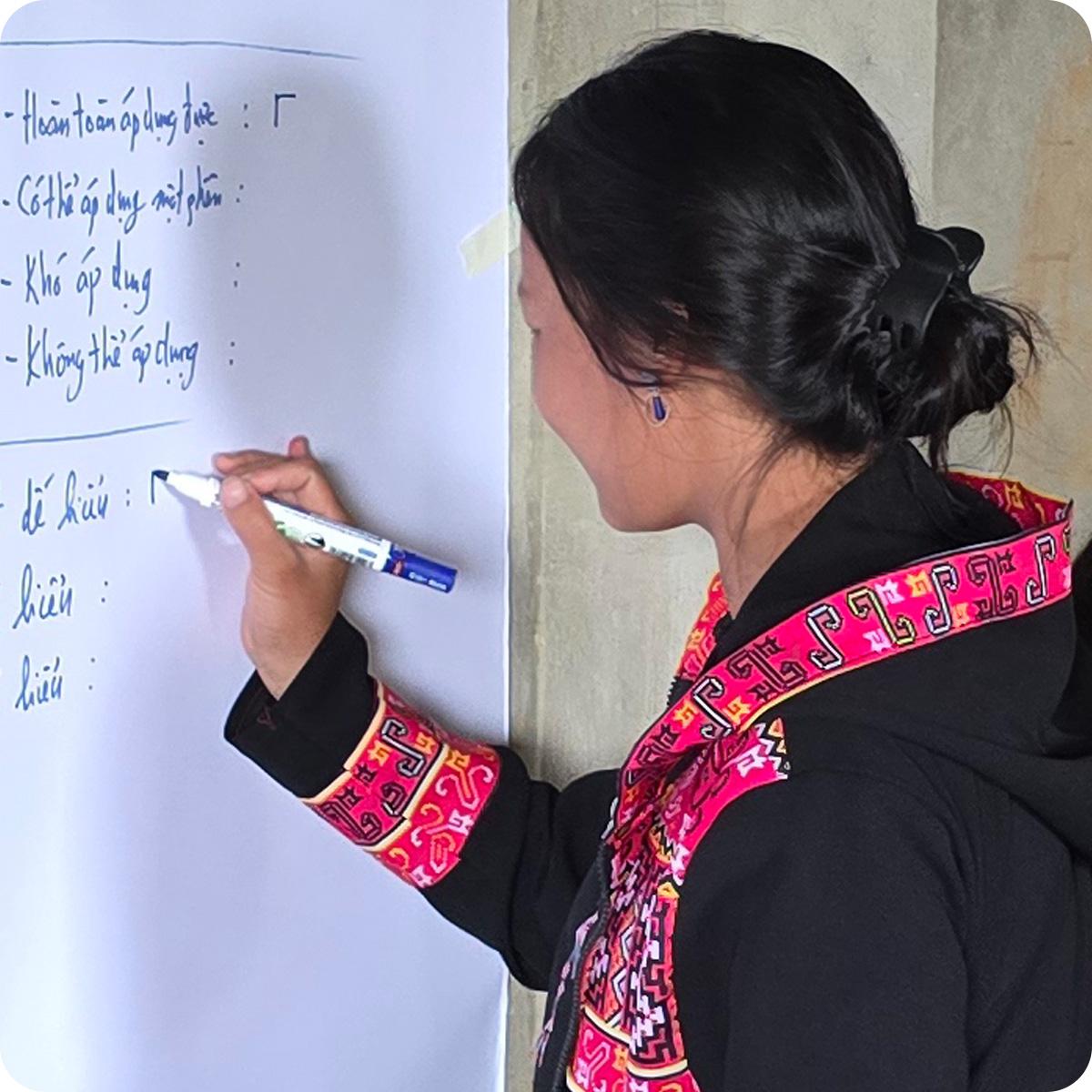Woman in black shirt with red embroidered trim writing on whiteboard. 
