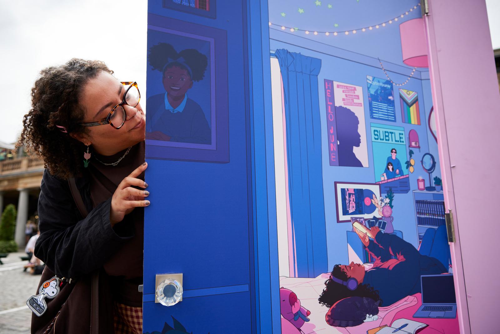 Woman in a black shirt with curly hair and glasses peeking around an opened door with an illustration on it.