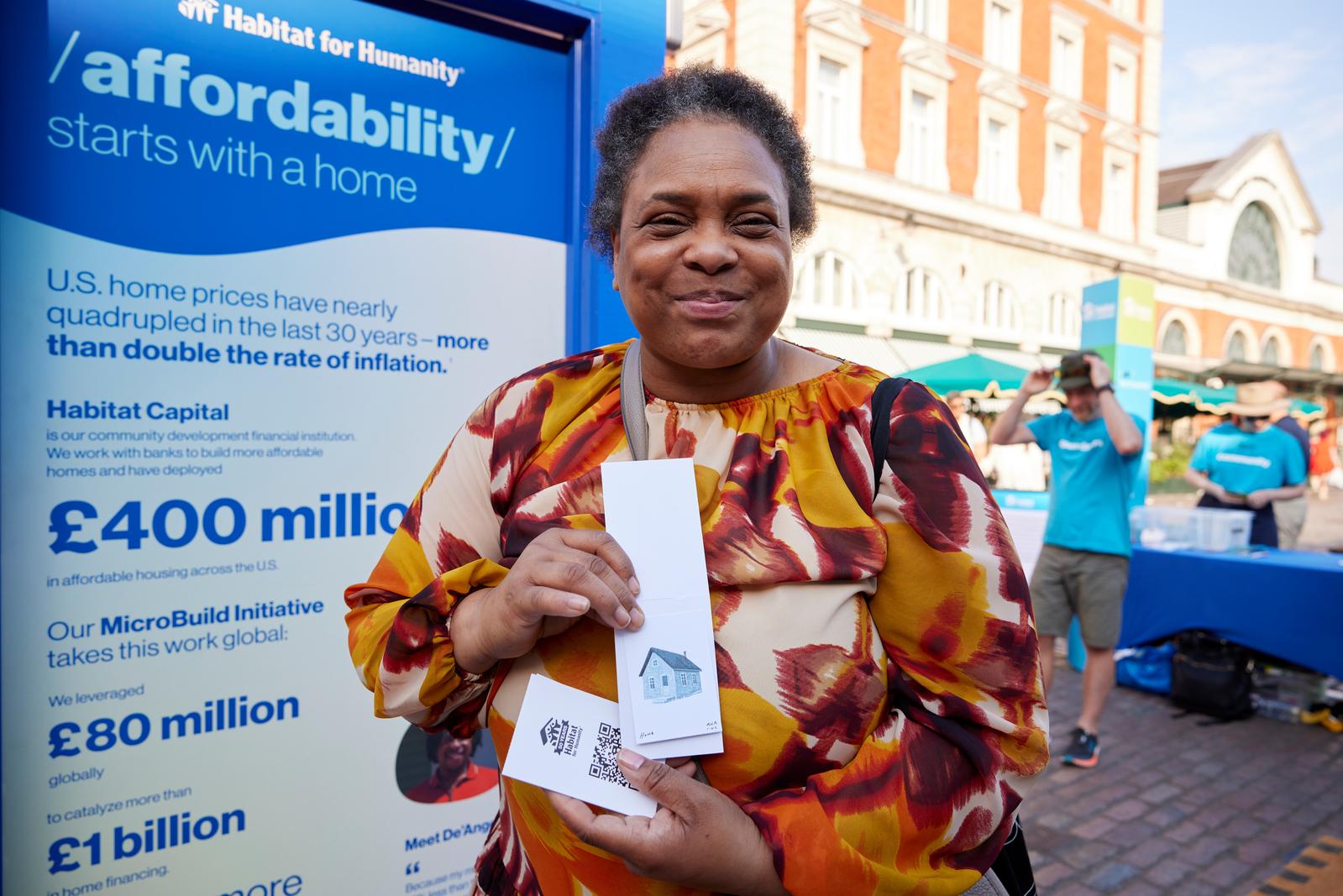 A woman in a multicolored brown shirt posing in front of a blue door discussing affordability holding up a mini house print.