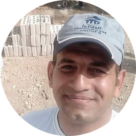 Fadi is wearing a Habitat baseball hat and he smiles for the camera. Behind him is a build site with blocks stacked on top of each other.