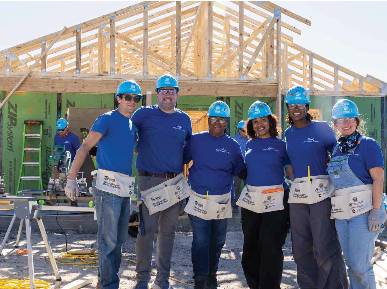A group of volunteers in blue Ford shirts wave at the camera while wearing blue Habitat hard hats.
