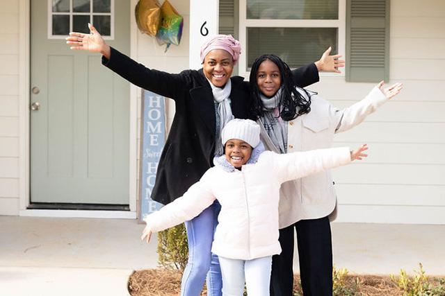 A woman and her two daughters pose with arms spread joyfully. They stand in the front entrance of their Habitat house with small but growing shrubs visible at their feet.