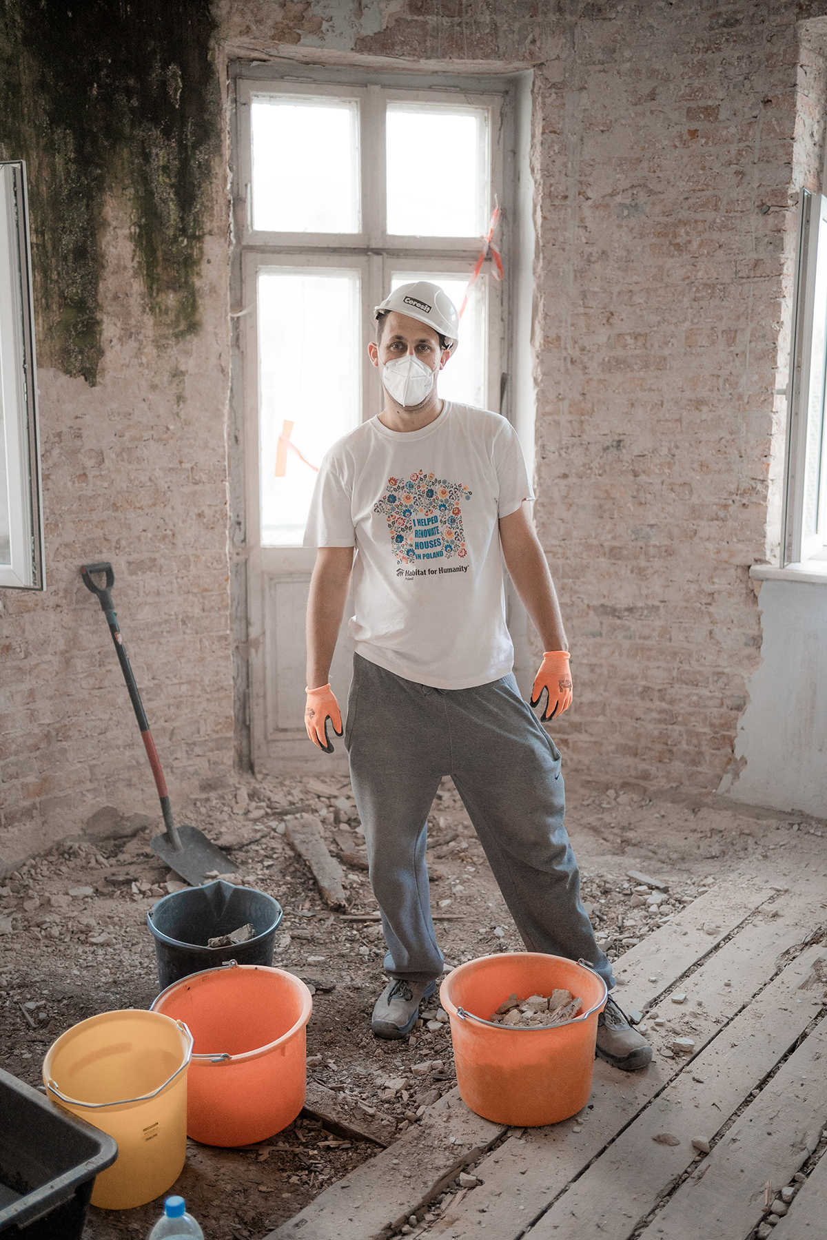 Lukasz, a volunteer at the Piaseczno build, stands in gray pants and a white t-shirt, hard hat, and face mask in front of three orange buckets in a build site.