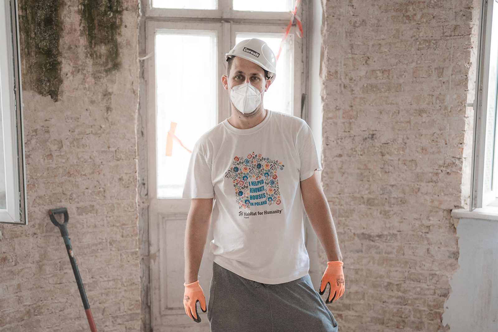 A volunteer stands in a white hard hat, mask, and gloves on a job site with a brick wall behind him.