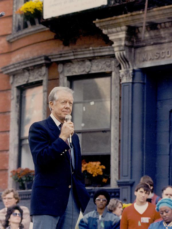 Jimmy Carter speaking to a crowd; he's wearing a navy suit and holding a microphone with a brick
