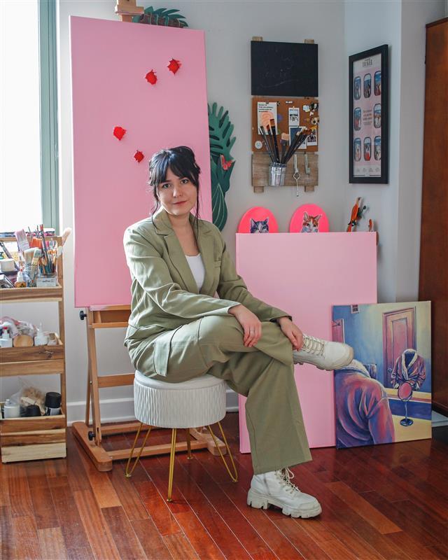 Woman in a tan suit and white boots sitting in front of a pink canvas and art supplies.