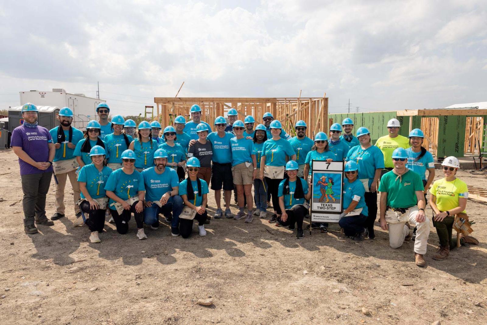 House seven volunteers stand in a large group in front of the frame of a house at Carter Work Project in Austin, Texas.