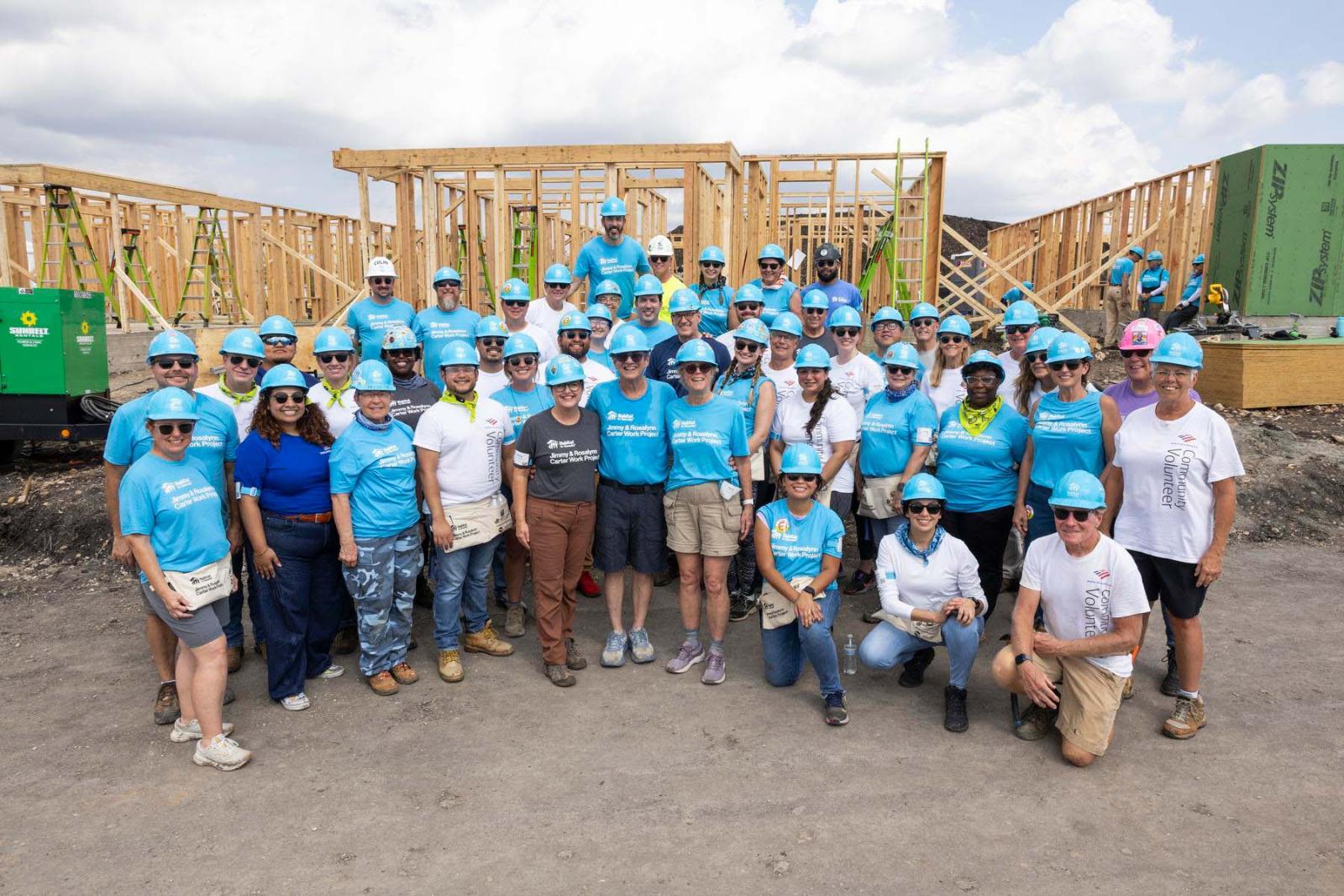 House twelve volunteers stand in a large group in front of the frame of a house at Carter Work Project in Austin, Texas.