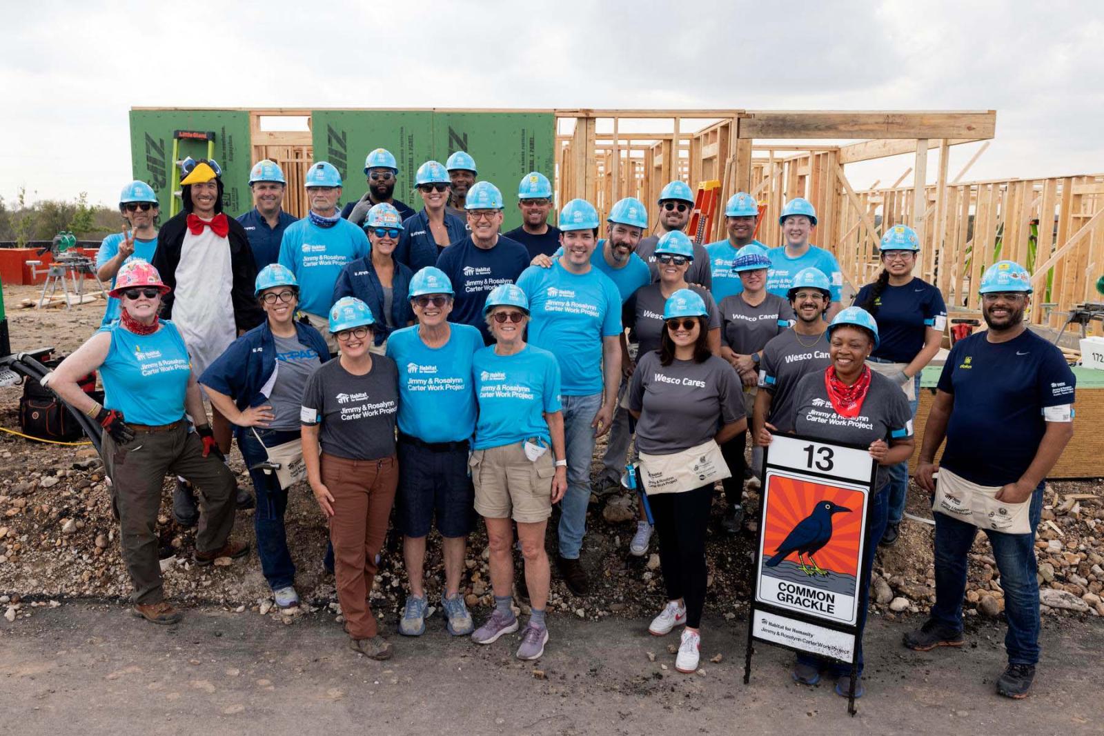 House thirteen volunteers stand in a large group in front of the frame of a house at Carter Work Project in Austin, Texas.