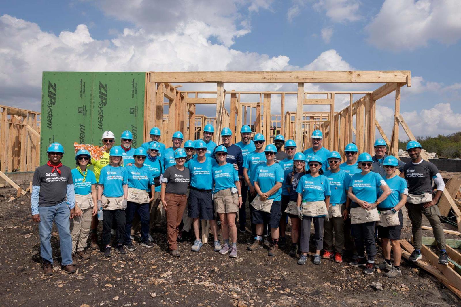 House fourteen volunteers stand in a large group in front of the frame of a house at Carter Work Project in Austin, Texas.