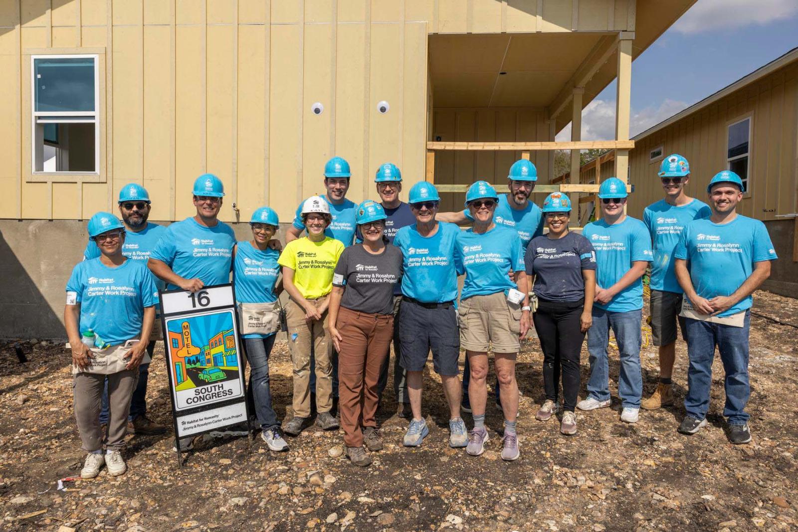 House sixteen volunteers stand in a large group in front of the frame of a house at Carter Work Project in Austin, Texas.
