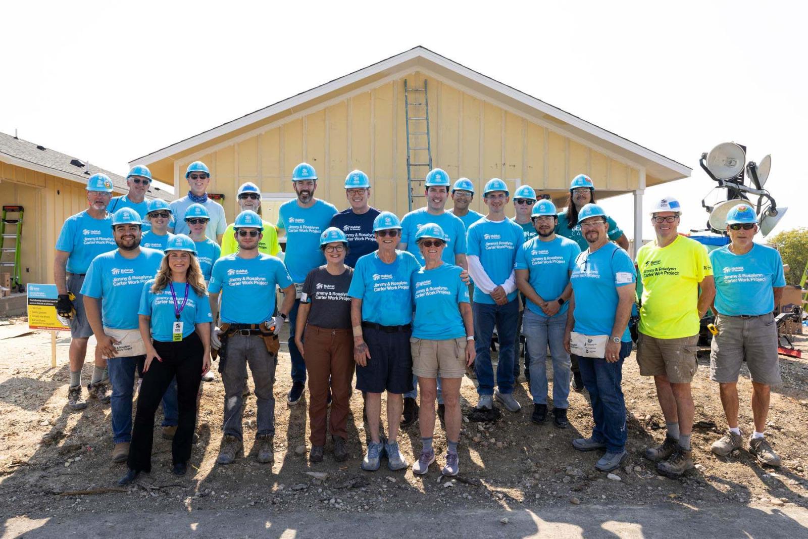 House seventeen volunteers stand in a large group in front of the frame of a house at Carter Work Project in Austin, Texas.