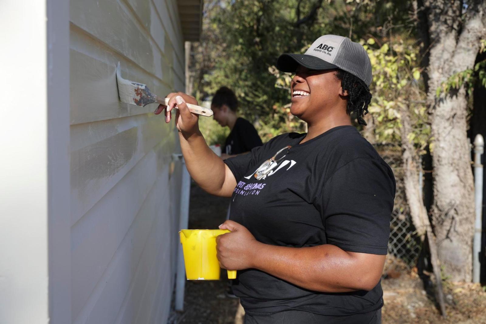 A volunteer holds a yellow cup and paints the outside wall of a house.
