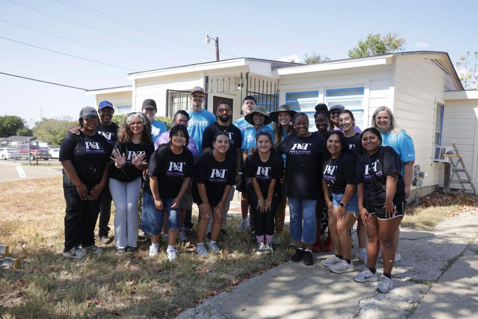 A group of volunteers in some Habitat shirts and some Peter and Thomas Foundation shirts stand in a semi-circle and pose in front of a home build.