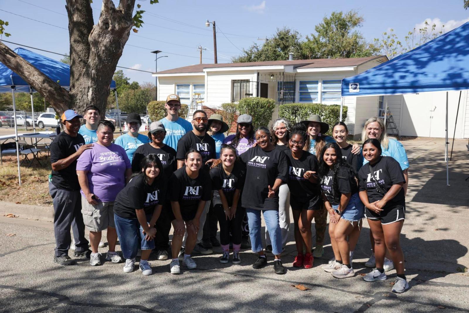 A group of volunteers in some Habitat shirts and some Peter and Thomas Foundation shirts stand in a semi-circle and pose in front of a home build.
