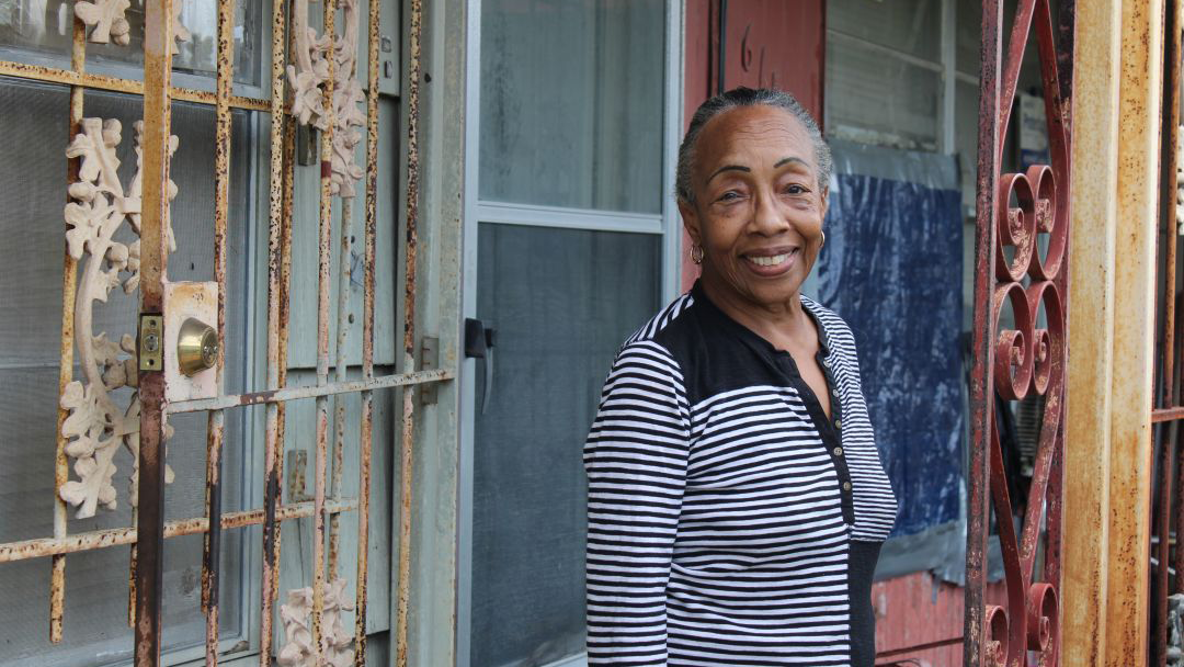 An elderly woman stands in front of the front door of her home and smiles.