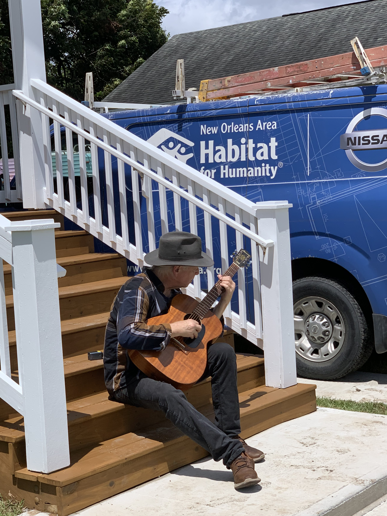 Man playing guitar on steps of house. Behind the steps a blue work van is visible. It displays the logos of Habitat for Humanity and Nissan