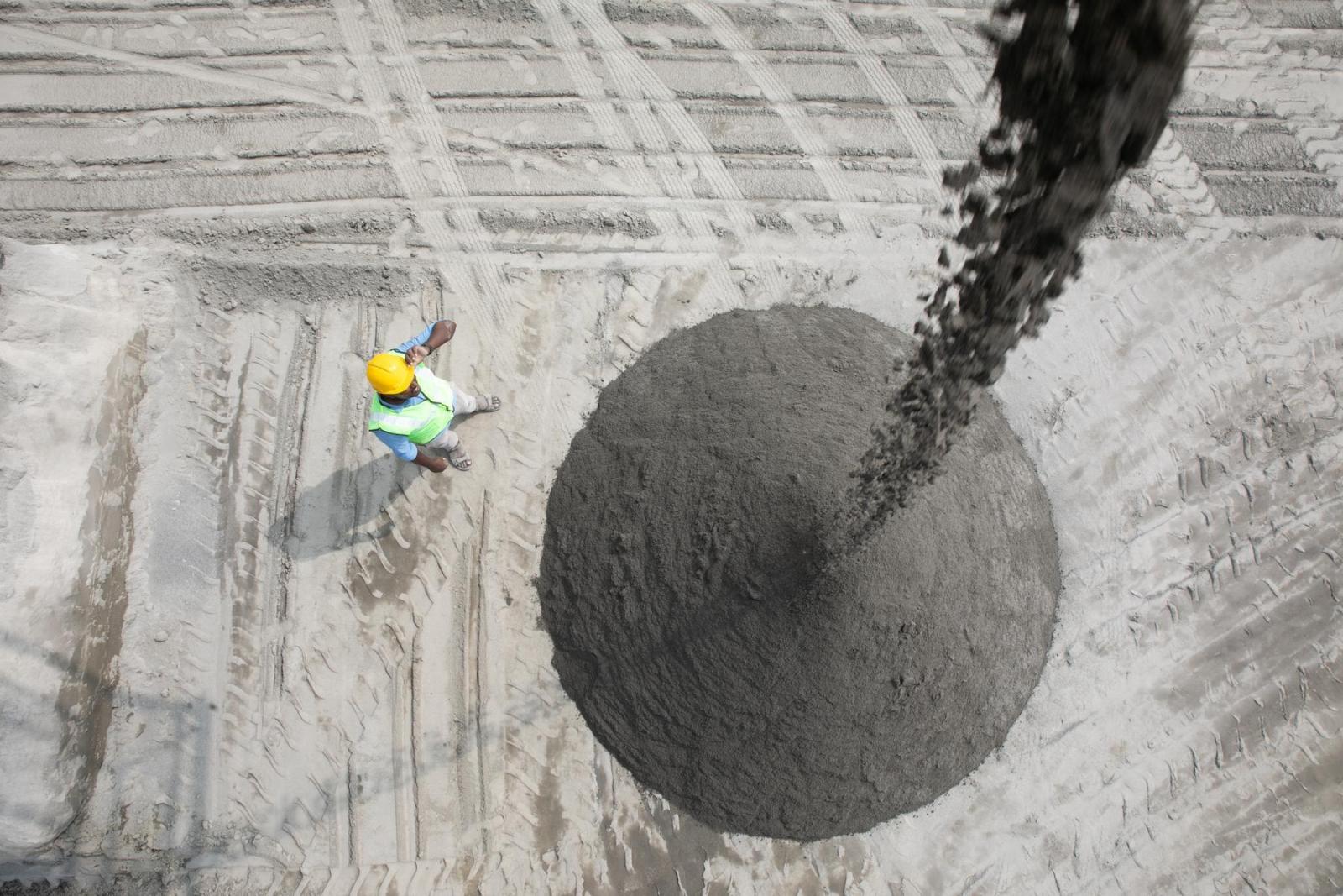 A man in a construction helmet stands on the ground below and watches while a stream of wet concrete streams down from above in a pile in front of him.
