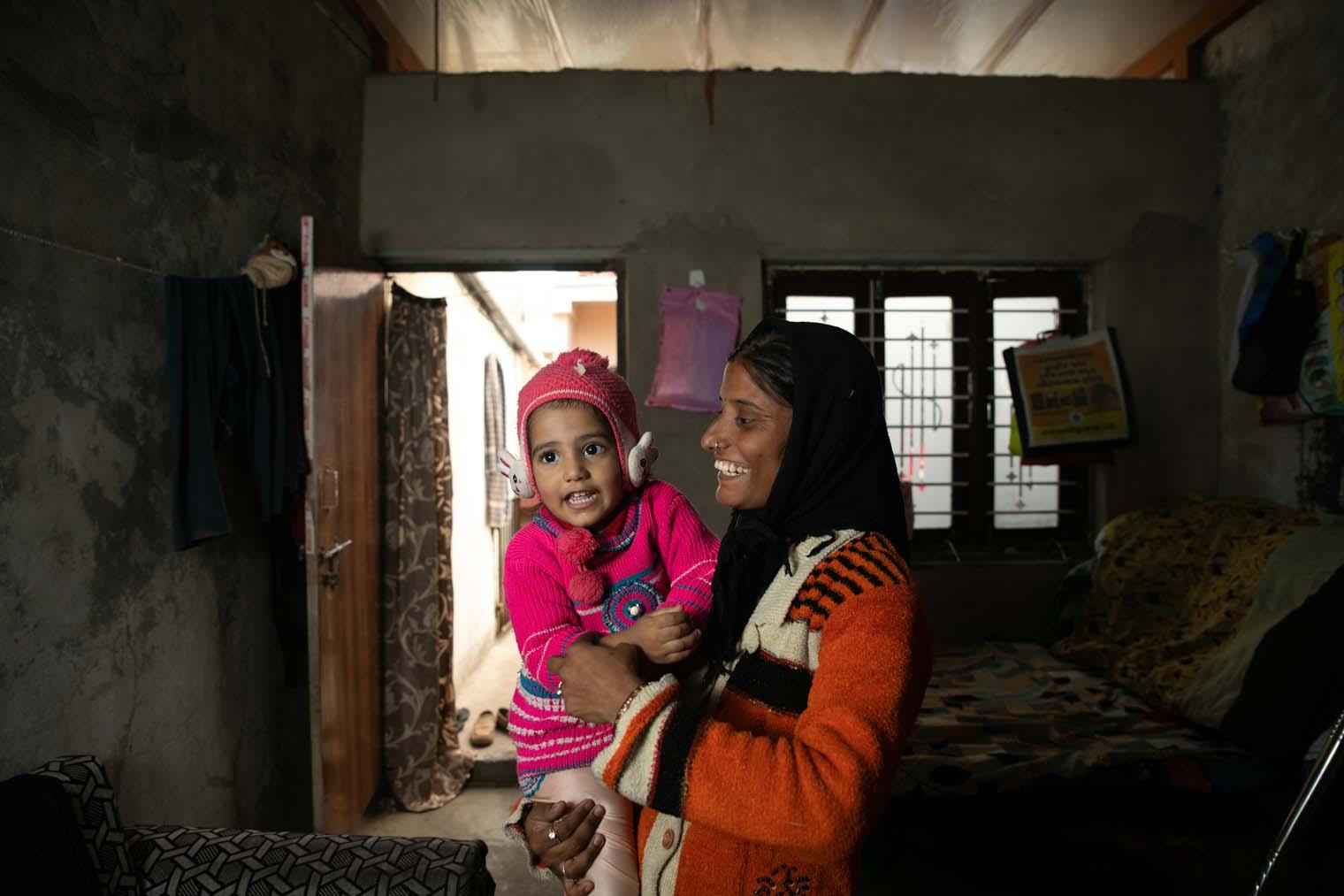 A woman in a sari stands inside her home and smiles at her daughter as she holds her up.