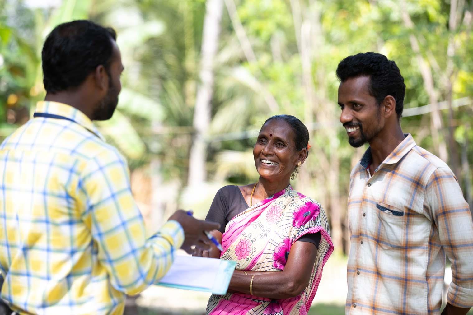 A man in a yellow plaid shirt stands and speaks to a couple while holding paperwork.