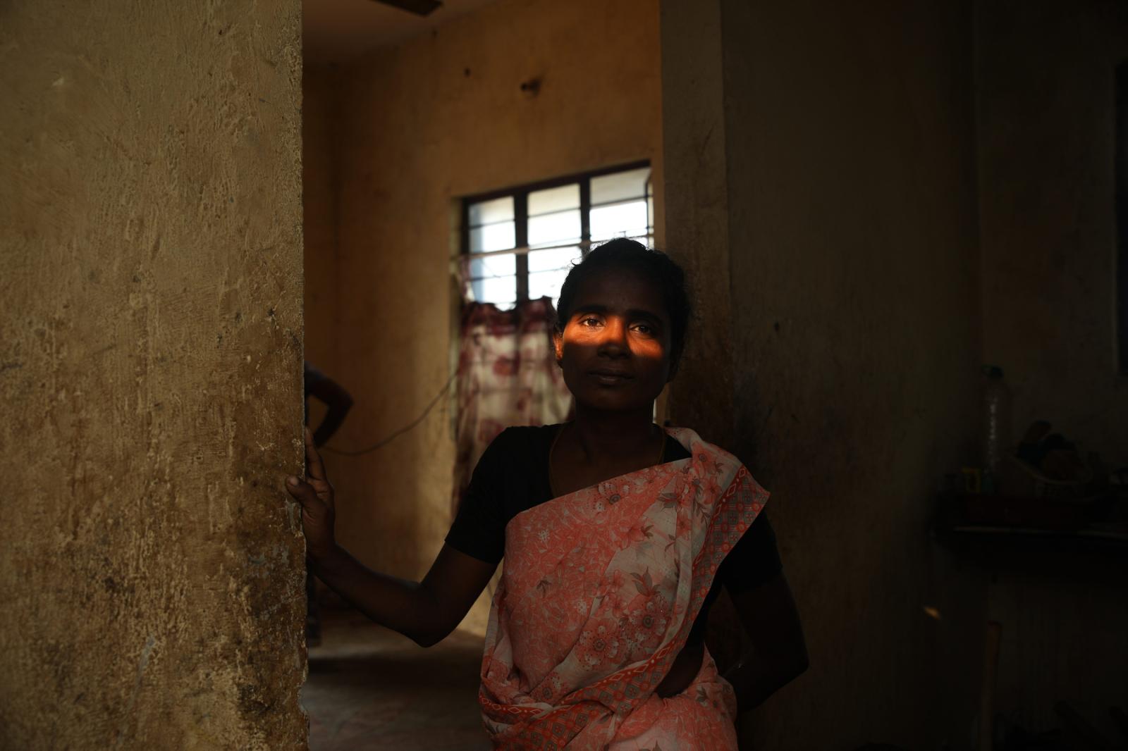 A woman in a sari stands in the shadows of her home. The light from the sun streams in through the window and across her eyes.