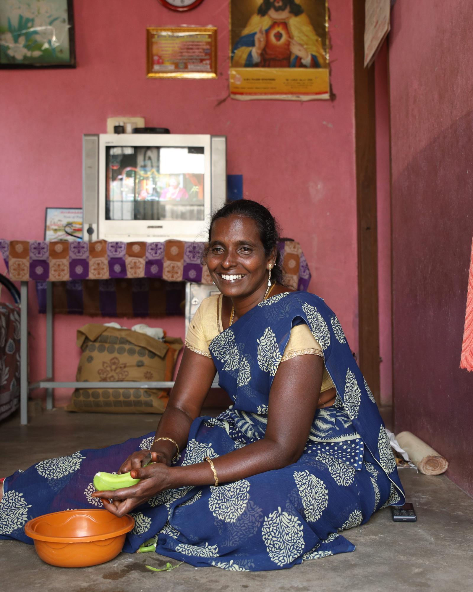 A woman sits on the floor of her home smiling as she peels a cucumber into a bowl.