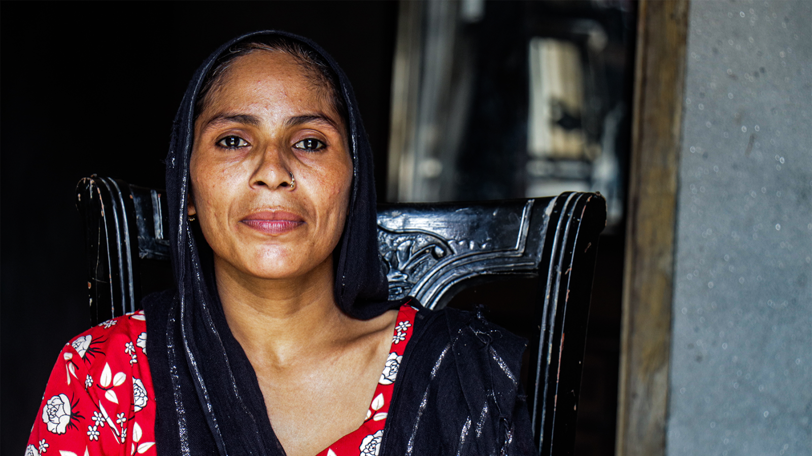 A woman in a black head scarf and red shirt sits in the doorway of her home, gazing at the camera.