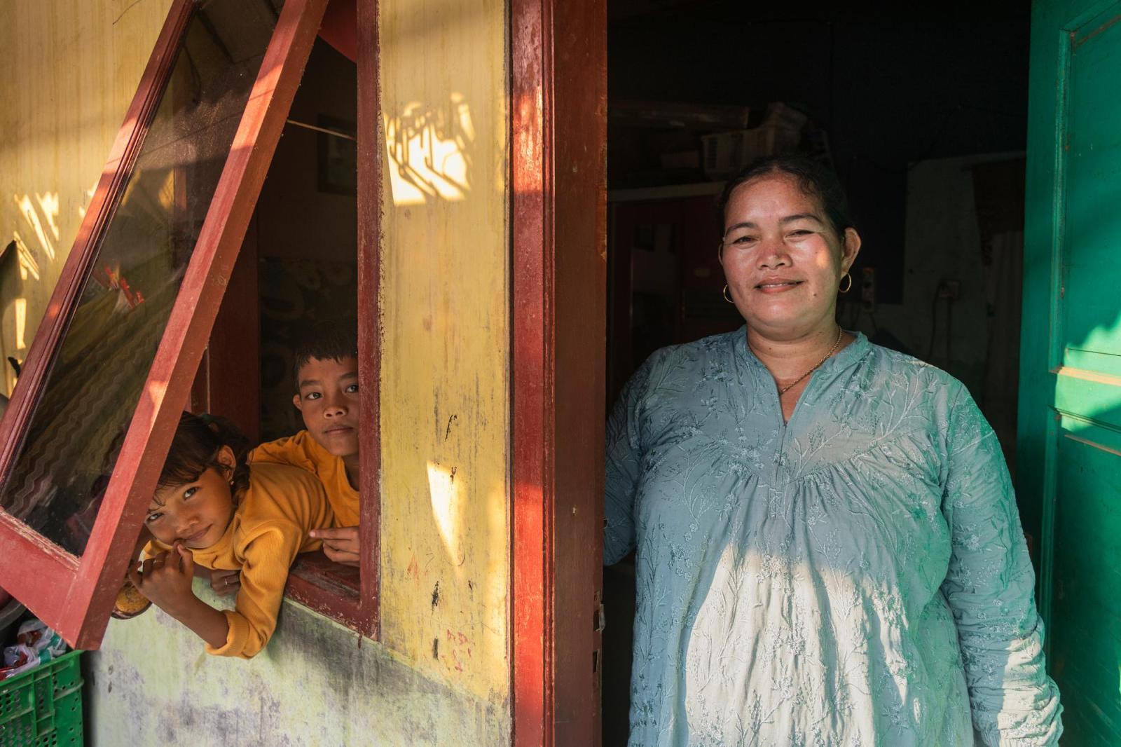 A smiling woman in a light blue embroidered shirt stands in her doorway. Her two children peek out of the open window.