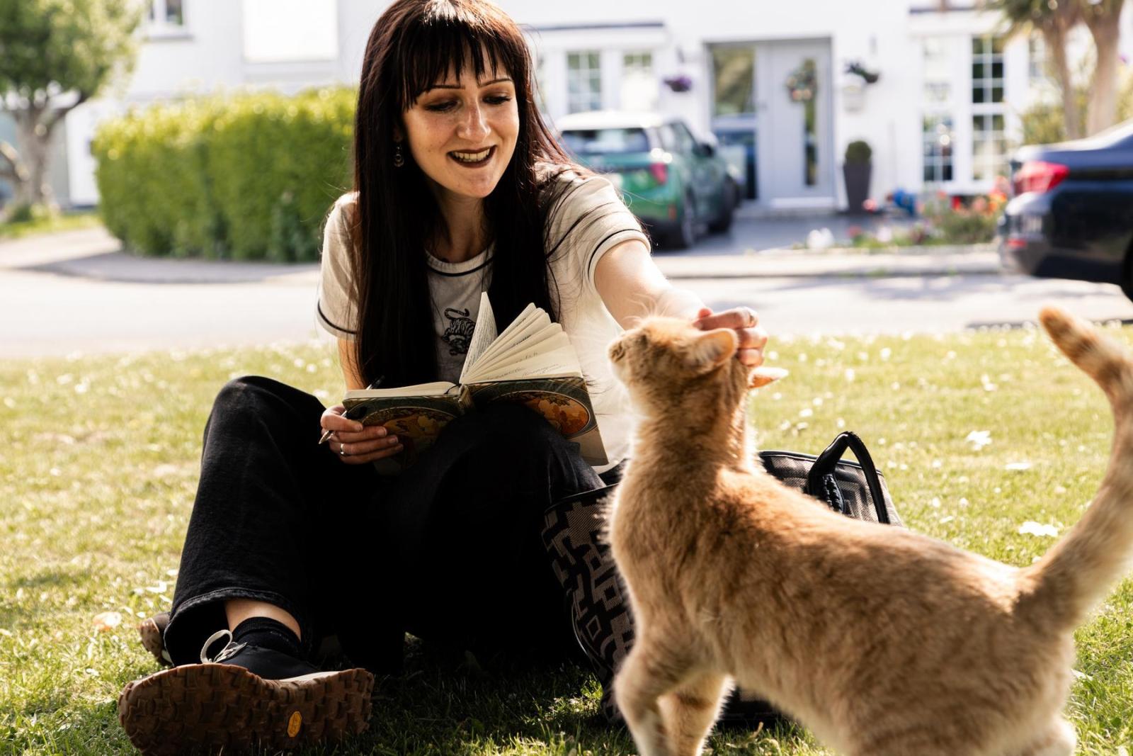A woman with long dark hair sits on grass holding a book and she reaches out to pet an orange cat.
