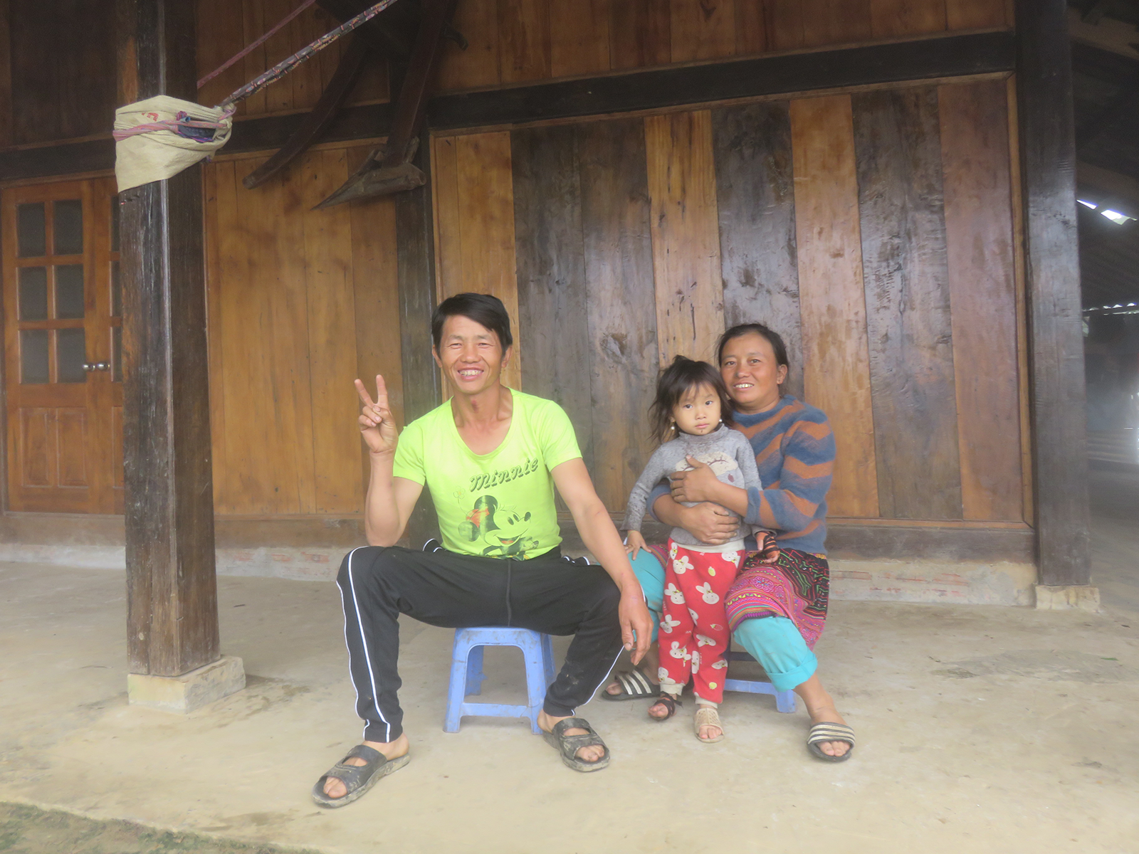 Smiling man seated on stool holding up two fingers in peace sign. Beside him, a smiling woman also seated on a stool hugs a small child. 