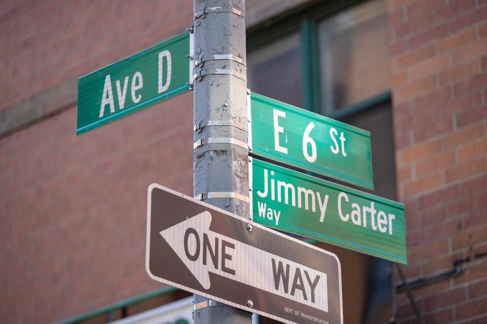 Street sign pole. Green street sign at top left reads "Ave D." On the right, green street signs read "E 6 St" and "Jimmy Carter Way." Below the other signs, a black and white arrow sign reads "ONE WAY."."