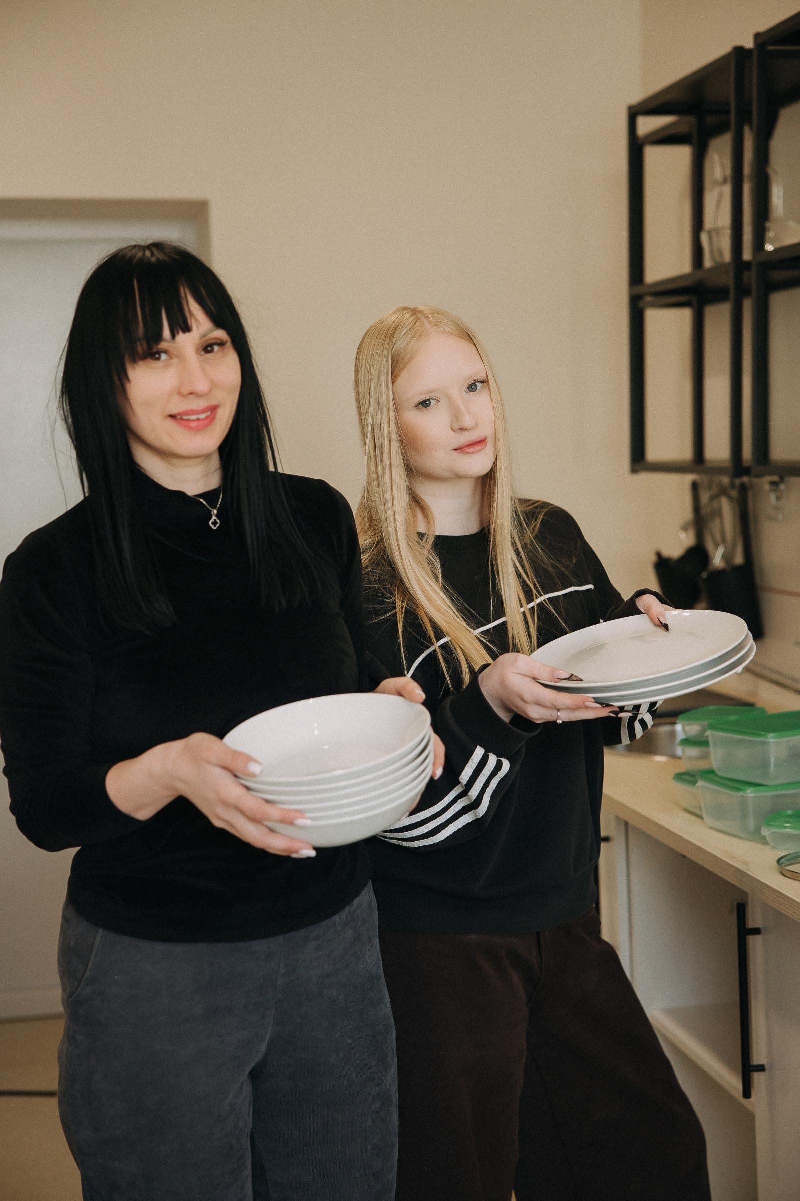 A woman and her teen daughter stand in their newly renovated kitchen holding stacks of plates and stare at the camera.