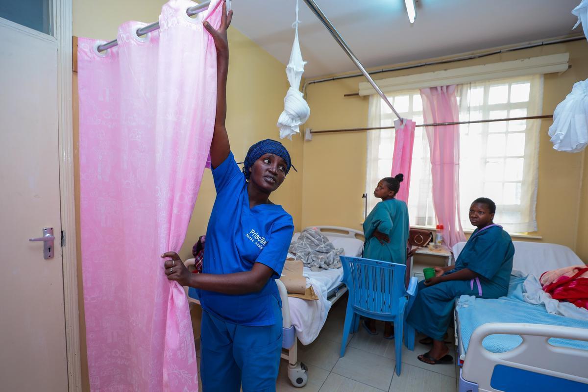 Kenyan nurse dressed in blue scrubs closing a pink curtain to maintain privacy for two other woman patients.
