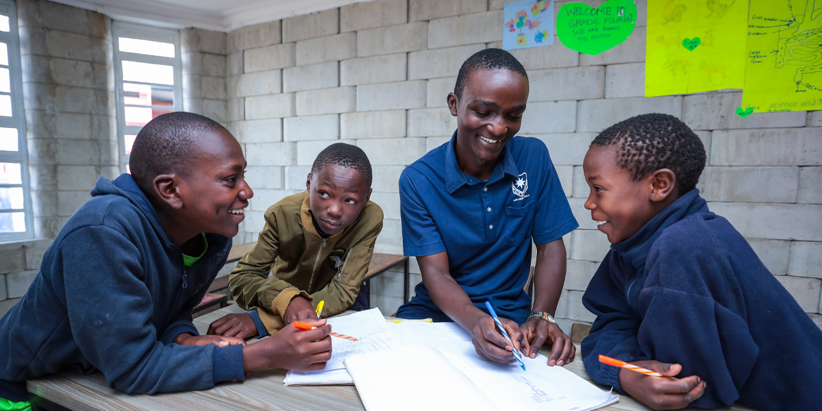 Four boys gather around a school table with pens and paper. Behind them, hand-written and drawn papers hang on the wall.