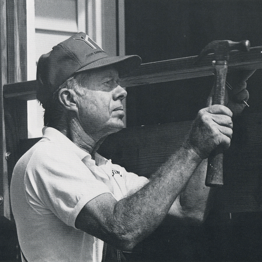 A black and white image of President Jimmy Carter wearing a baseball hat and holding a hammer to a beam on a Habitat worksite.