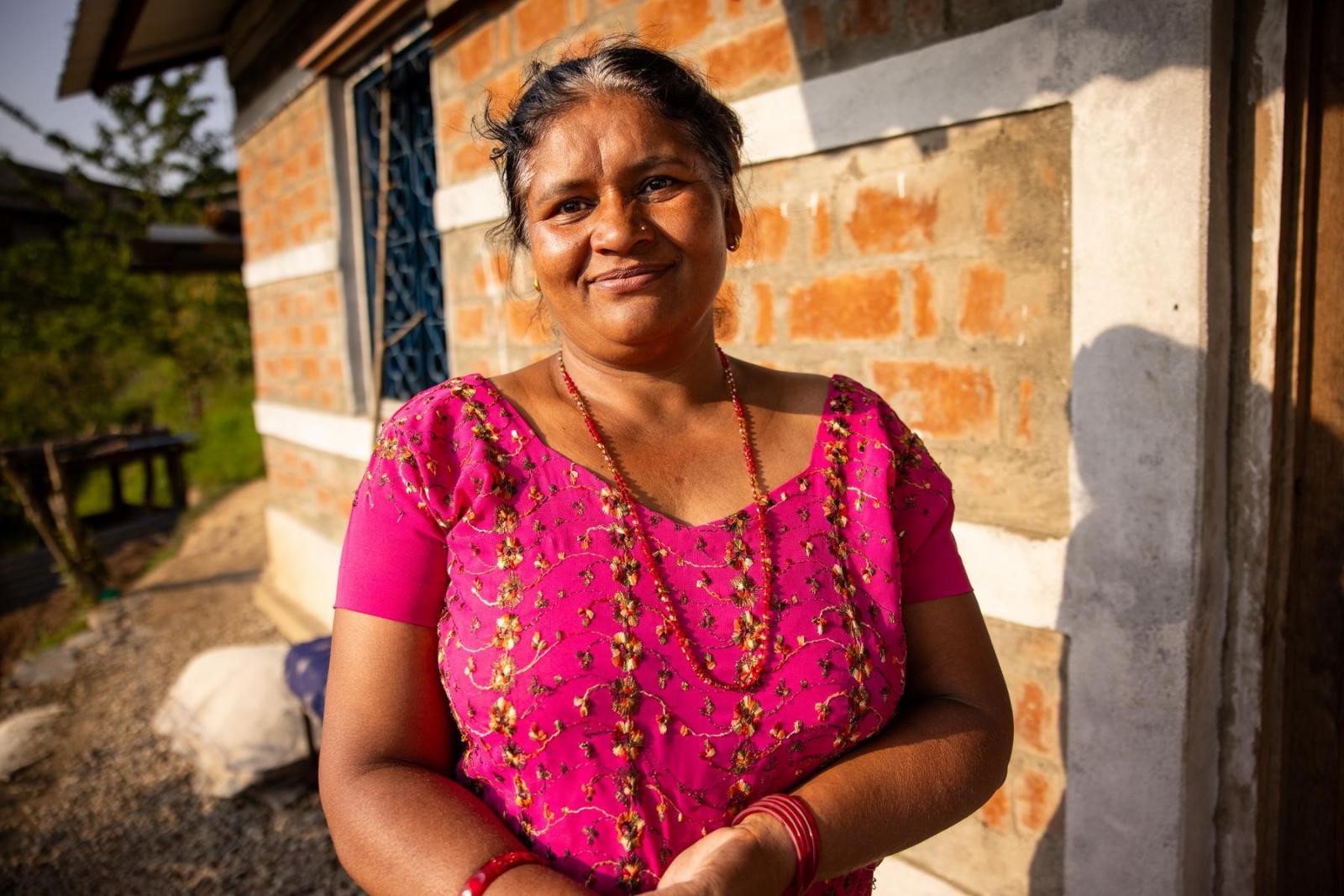 Niru stands right outside the front door of her brick home. She is wearing a pink sari and necklace and smiles as the sun sets.
