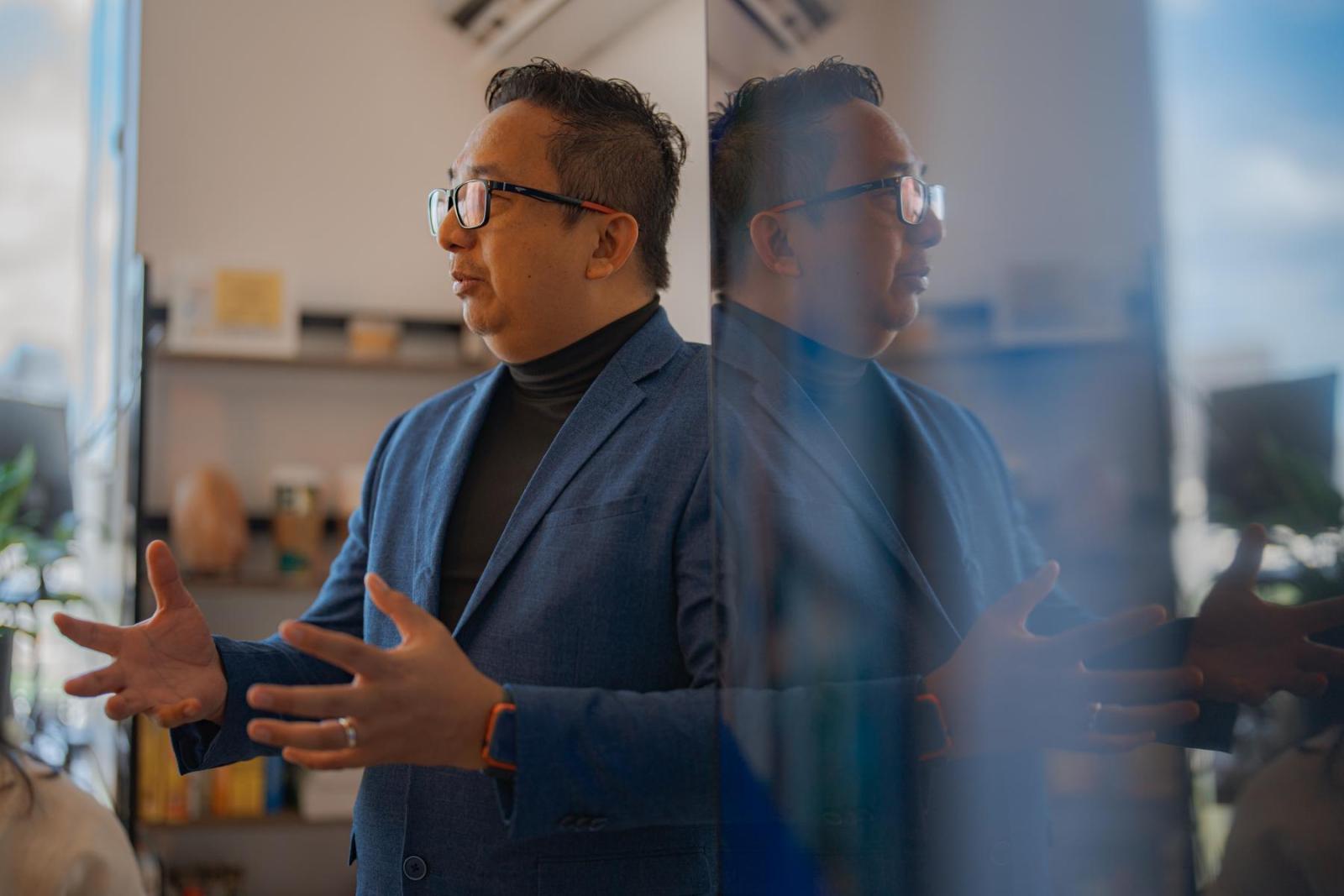 A man stands in a blue blazer and presents to an audience. His reflection can be seen in glass next to him.
