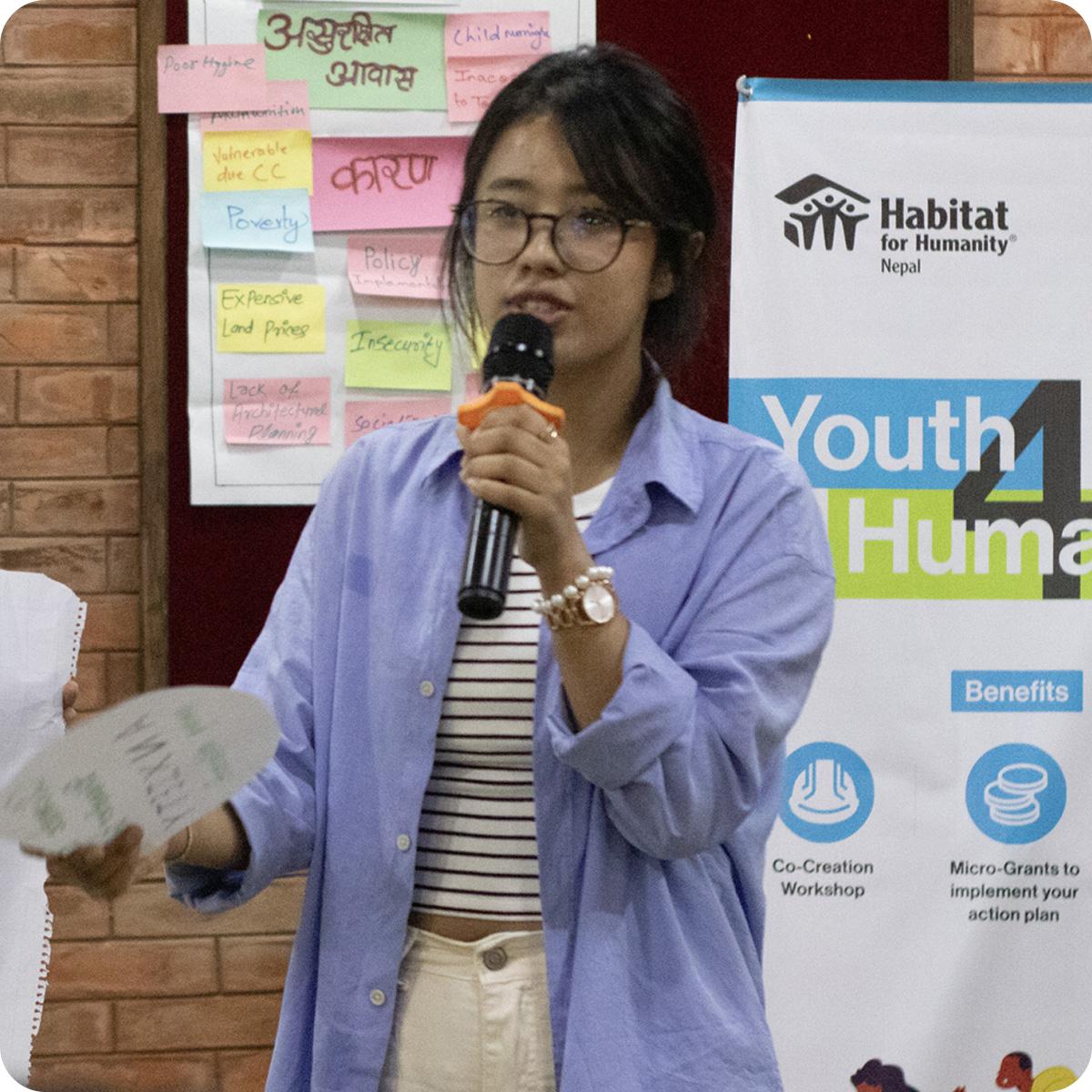 Woman in blue shirt speaking into microphone. Behind her is a banner that says "Youth4Humanity."