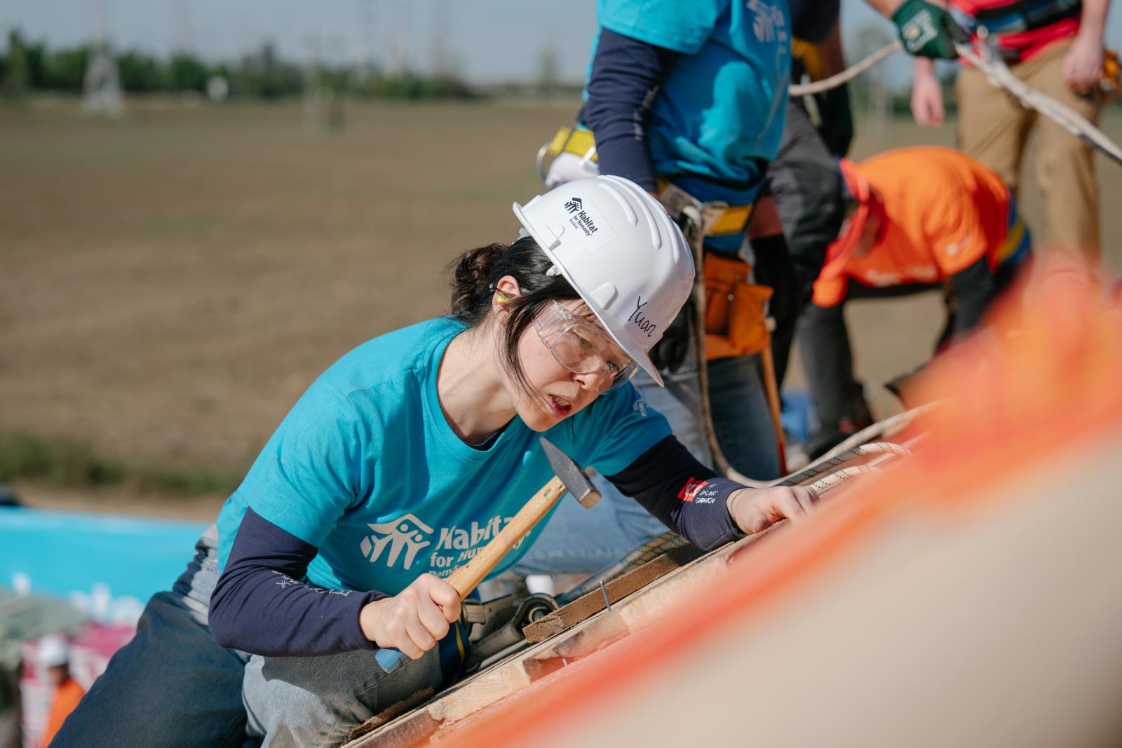 Volunteer in a blue Habitat shirt and a white hard hat hammering.