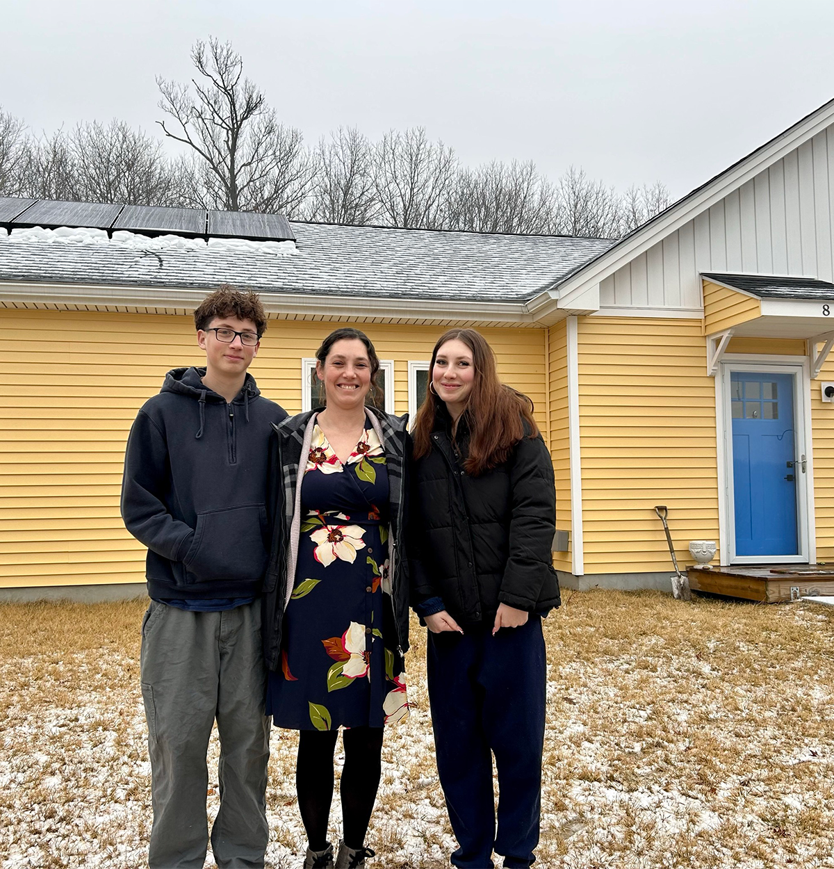 A woman stands between her two children in front of her yellow home that has solar panels on the roof.
