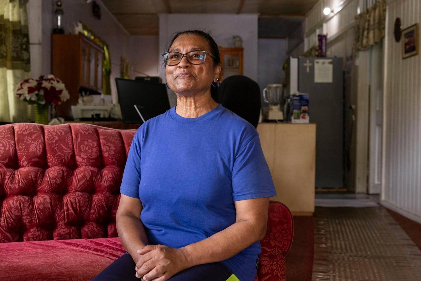 A woman in a royal blue t-shirts sits with her arms folded in her lap on a red tufted couch.